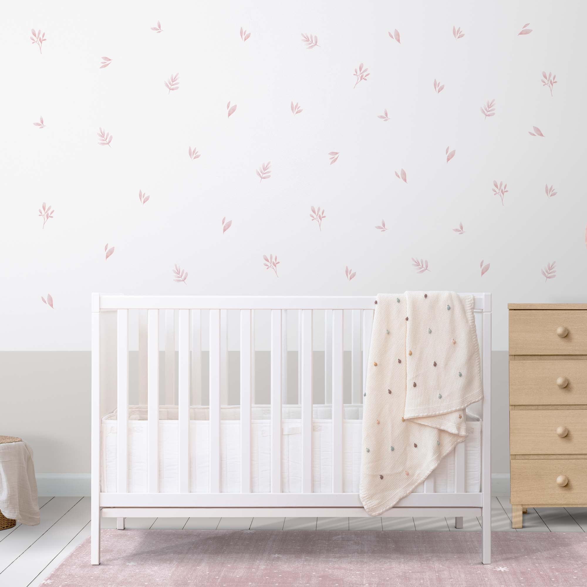 Vista general de habitación de bebé con vinilos de plantas en tonos rosados aplicados sobre pared blanca, creando un ambiente natural y acogedor. Cuna blanca en habitación de bebé con pared decorada con vinilos de hojas en acuarela rosa, simulando papel pintado botánico #color_plantas-acuarela-rosa